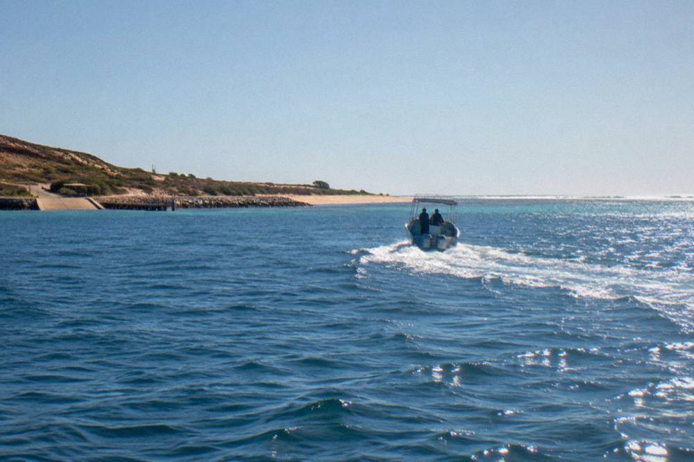 A small charter boat crossing open water toward a low reef line at midday