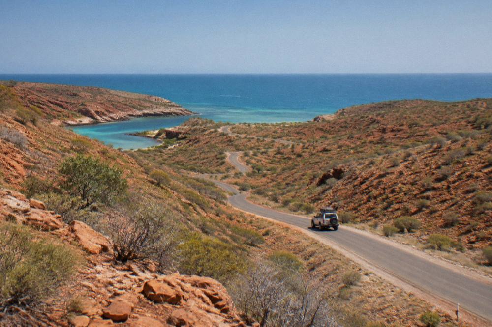 A sealed road winding through red rock hills toward a distant blue ocean