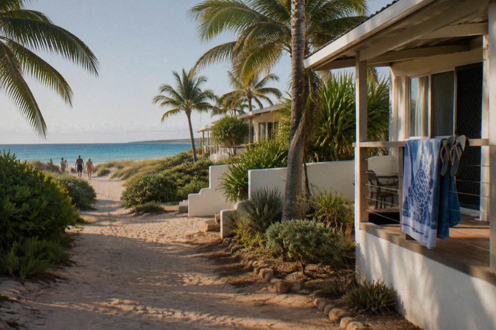 Palm-fronted accommodation and a sandy path leading toward the Coral Bay beachfront