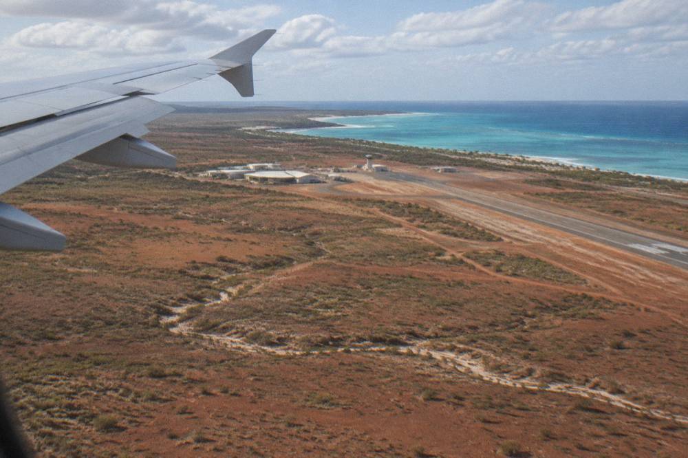 Aircraft wing above red outback plains and a pale coastline approaching Learmonth